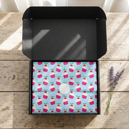 Open black SP Boutique gift box with light blue birthday tissue paper printed with pink cupcakes and balloons, folded and sealed with a round white sticker
